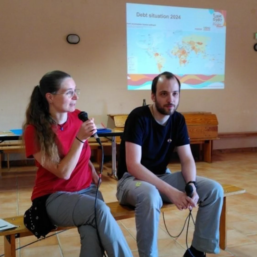 Das Bild zeigt zwei Personen in einem Saal, die auf einer Bank sitzen und ein Mikrofon halten. Im Hintergrund ist das Bild einer Präsentation sichtbar.