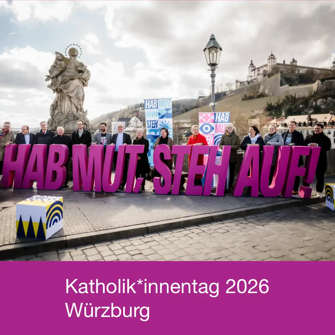 Das Bild zeigt vor der Kulisse Würzburgs im Hintergrund, Menschen, die in einer Reihe auf einer Brücke stehen. Vor den menschen stehen meterhohe Buchstaben, die das Motto des Kirchentages bilden: Hab Mut, steh auf! Weiterer Text: "Katholik*innentag 2026 Würzburg".