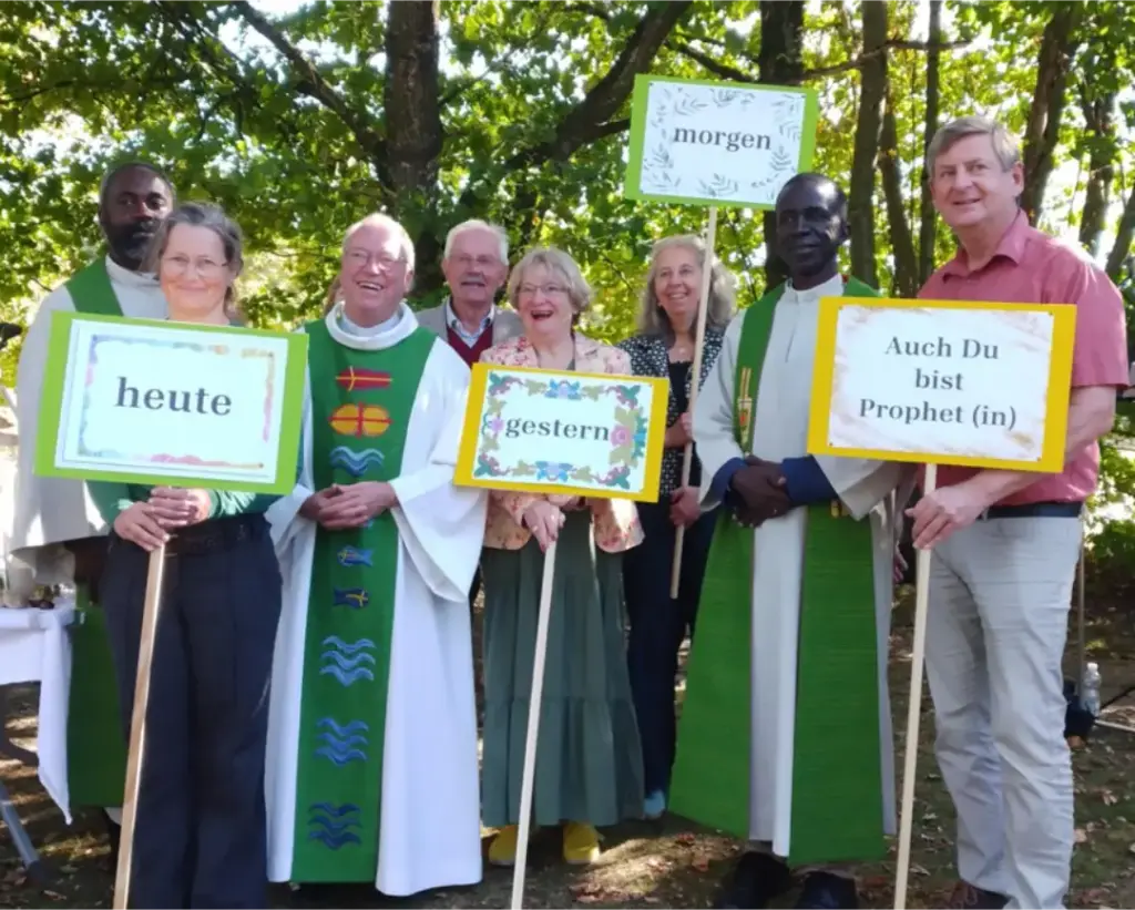 Das Bild zeigt ein Gruppenbild mit acht Menschen, die in einer Reihe stehen. Vier Schilder sind sichtbar. Aufschriften: "heute" "Gestern" "morgen" und "Auch Du bist Prophet(in)".